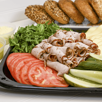 A bagel and topping tray with deli meats, sliced tomatoes, pickles, and greens