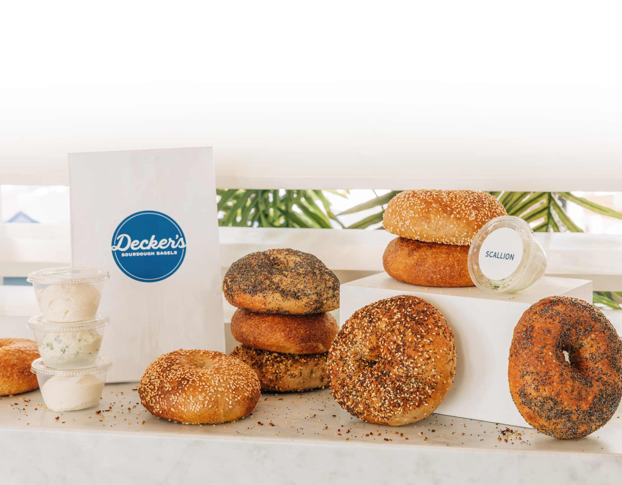 A variety of bagels stacked on a white counter in front of a white bag with a Decker logo printed on it.